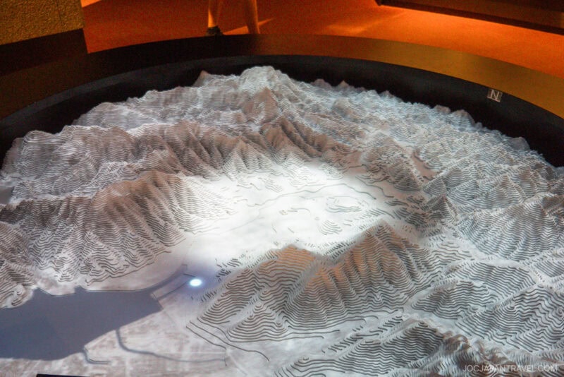 A detailed 3D topographic model displays a mountainous landscape with contour lines, showing valleys and peaks under soft lighting in the Nagasaki Atomic Bomb Museum exhibition setting.