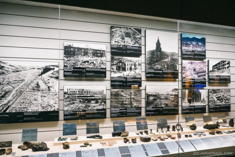 The Nagasaki Atomic Bomb Museum exhibit displays black-and-white photos of war devastation, with captions, above a collection of artifacts and informational cards presented behind glass.