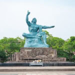 Nagasaki Atomic Bomb Museum and Peace Park (長崎原爆資料館・平和公園) A large bronze statue of a seated man with one arm raised and the other extended sits on a stone pedestal near the Nagasaki Atomic Bomb Museum, surrounded by greenery and a paved plaza. Flowers are placed at the base of the statue.