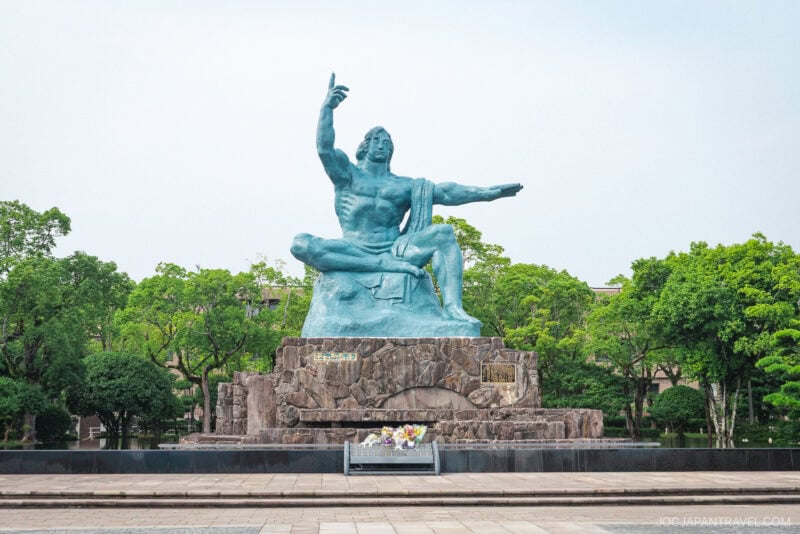 A large bronze statue of a seated man with one arm raised and the other extended sits on a stone pedestal near the Nagasaki Atomic Bomb Museum, surrounded by greenery and a paved plaza. Flowers are placed at the base of the statue.