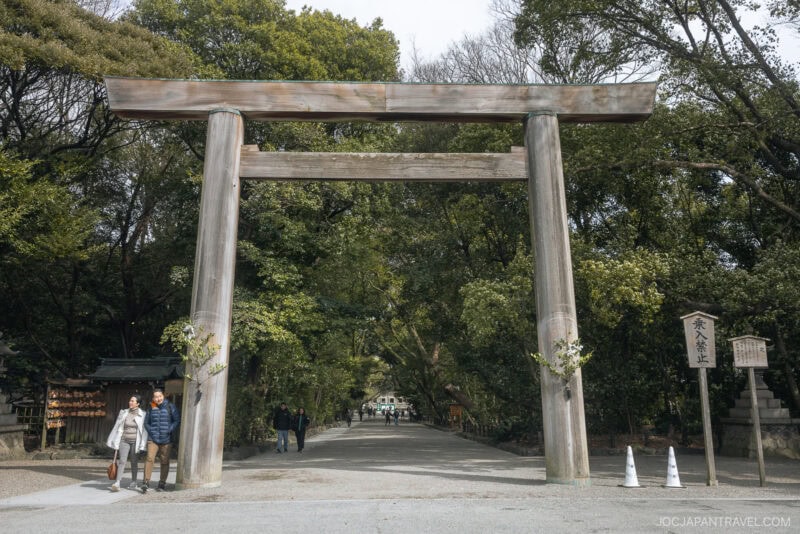A large wooden torii gate stands at the entrance of a tree-lined path at Atsuta Shrine. People walk beneath the gate, surrounded by lush greenery, while a wooden sign with Japanese writing stands to the right of the path.