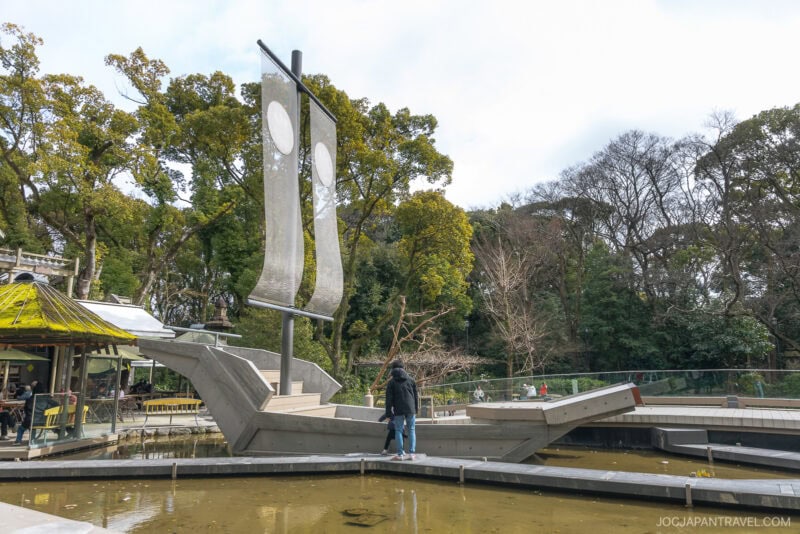 A person stands on a modern, concrete boat-shaped structure with tall sails in a shallow pond at Atsuta Shrine, surrounded by trees and a small building on the left. The scene appears to be part of the shrine’s scenic park grounds.
