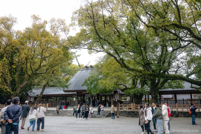 People visit Atsuta Shrine, a traditional Japanese shrine surrounded by large, leafy trees. The wooden structure with its sloped roof stands in an open courtyard where visitors walk, stand, and take photos.