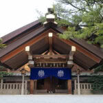 Atsuta Shrine (Atsuta Jingu): Discover Nagoya’s Most Sacred Site A traditional Japanese shrine, reminiscent of Atsuta Shrine, features a large wooden roof, blue curtain with white emblems, stone lanterns, and is surrounded by lush green trees.