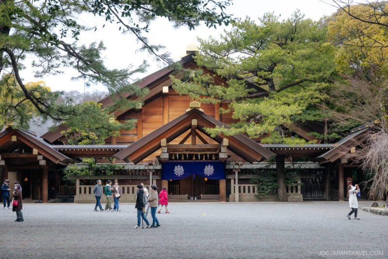 People walk and take photos in front of the grand wooden Atsuta Shrine surrounded by trees, with a gravel courtyard in the foreground.