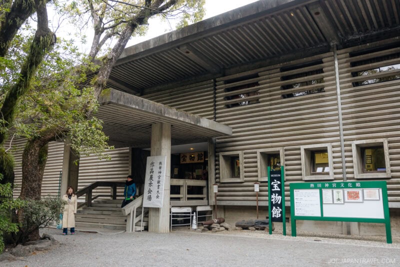 A modern concrete and wooden building with horizontal slats, signs in Japanese, a green information board, and two people standing near the entrance—possibly at Atsuta Shrine—surrounded by trees.
