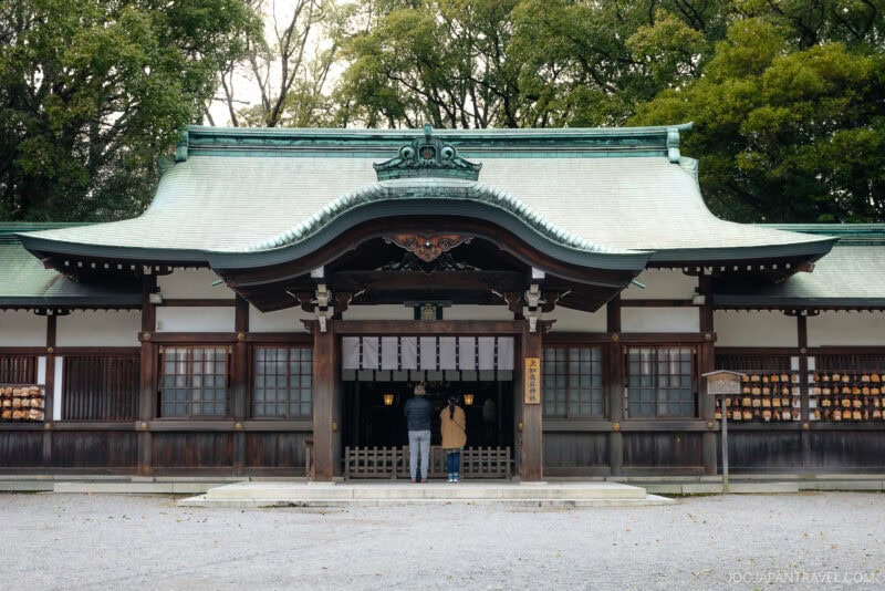 Two people stand in front of Atsuta Shrine, a traditional Japanese shrine with a green curved roof, wooden beams, and paper lanterns on both sides. Tall trees rise in the background.