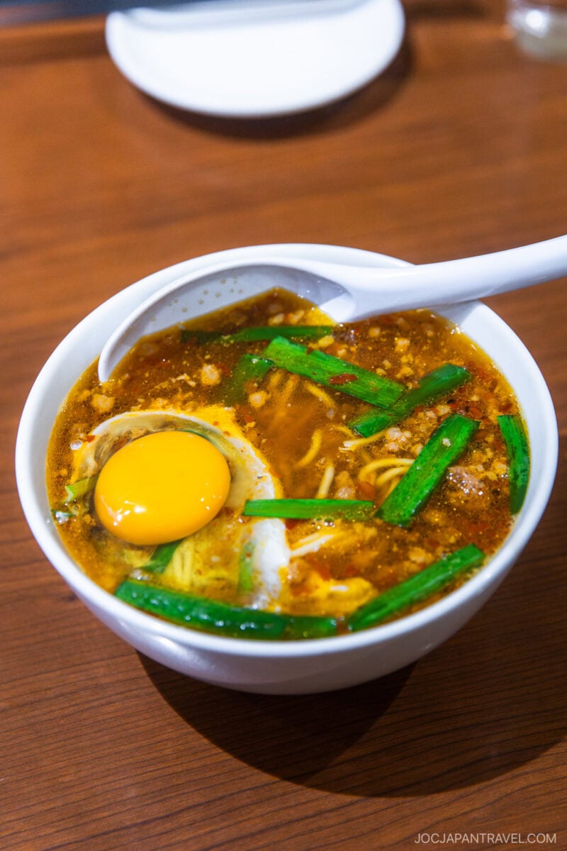 A bowl of ramen with a raw egg yolk, sliced green onions, and rich broth, served with a white spoon on a wooden table—a perfect addition to any Nagoya food guide.