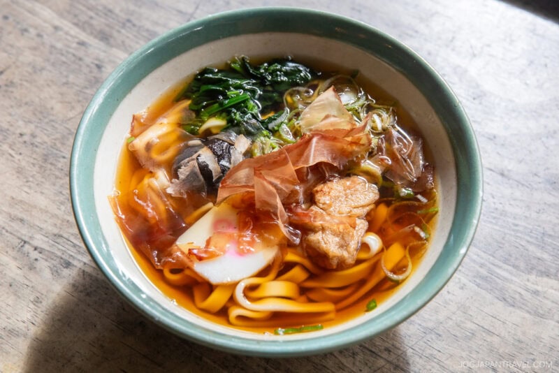 A bowl of udon noodle soup with sliced fish cake, greens, bonito flakes, broth, and chunks of meat is served in a green and white ceramic bowl on a wooden surface—a delicious highlight for any nagoya food guide.