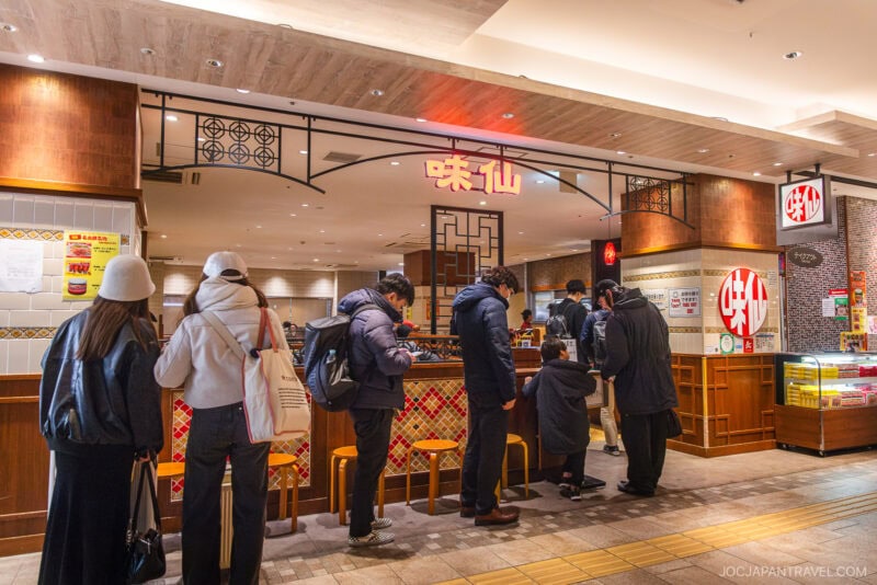 People wait in line at a brightly lit ramen restaurant featured in the Nagoya food guide. Some are seated at the counter eating, while others stand with bags and coats, facing the order counter.