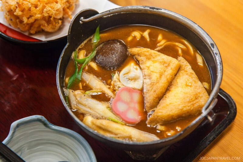 A bowl of Japanese miso udon soup topped with fried tofu triangles, sliced fish cake, shiitake mushroom, green onions, and vegetables—an enticing highlight featured in our Nagoya food guide—served on a wooden tray with sides nearby.