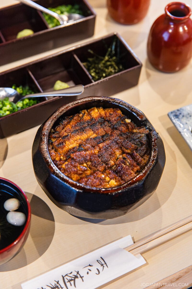 A bowl of grilled eel with rice (hitsumabushi) sits on a table, surrounded by condiments, garnish trays, chopsticks, and a red teapot—an essential dish featured in any Nagoya food guide for a traditional Japanese meal.