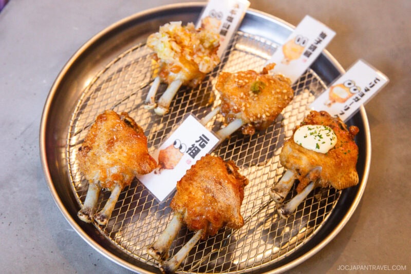 Six pieces of grilled chicken wings with exposed bones are arranged on a round metal tray, each with a different topping and a paper label—perfect for any nagoya food guide—set on a mesh wire rack.