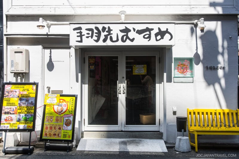 A small Japanese restaurant featured in the Nagoya food guide, with a white exterior, glass double doors, and a sign with Japanese characters above. A yellow bench and menu displays stand outside in the sunlight.