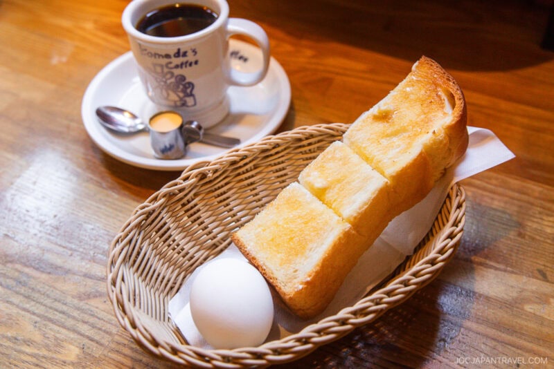 A basket with a slice of thick buttered toast and a hard-boiled egg, next to a cup of black coffee—classic items you’ll find in any Nagoya food guide—all set on a wooden table.