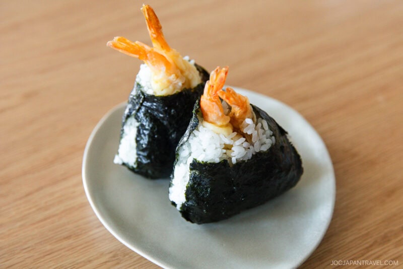 Two shrimp tempura onigiri, featured in our Nagoya food guide, are wrapped in seaweed and filled with white rice and crispy shrimp, sitting on a small round white plate atop a wooden table.