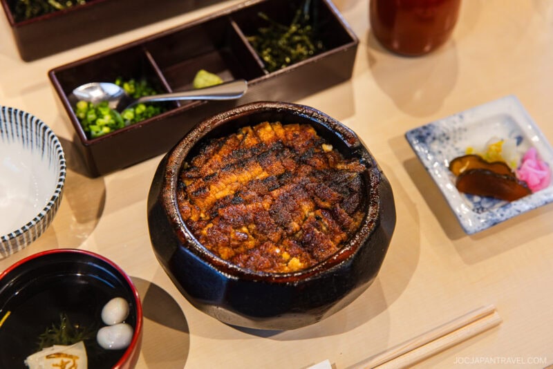 A bowl of grilled eel on rice (hitsumabushi), a highlight in any Nagoya food guide, is served with condiments, soup, pickles, and a tea set on a wooden table. Chopsticks, a small dish, and a drink are also visible.