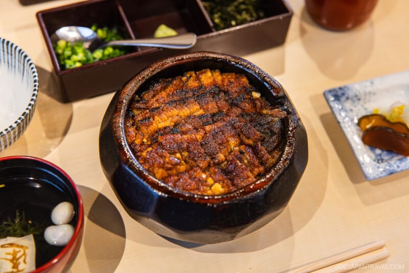 A bowl of grilled unagi (eel) over rice, served with sides of pickles, soup, and condiments on a light wooden table—a highlight featured in many Nagoya food guide recommendations.