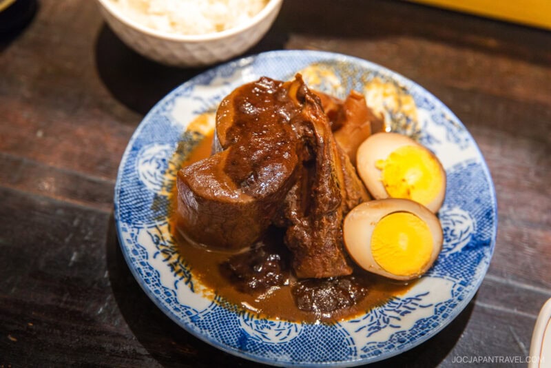 A blue and white plate with braised beef in a rich brown sauce, served with two halved boiled eggs; a bowl of white rice is visible in the background—a savory highlight from any Nagoya food guide.