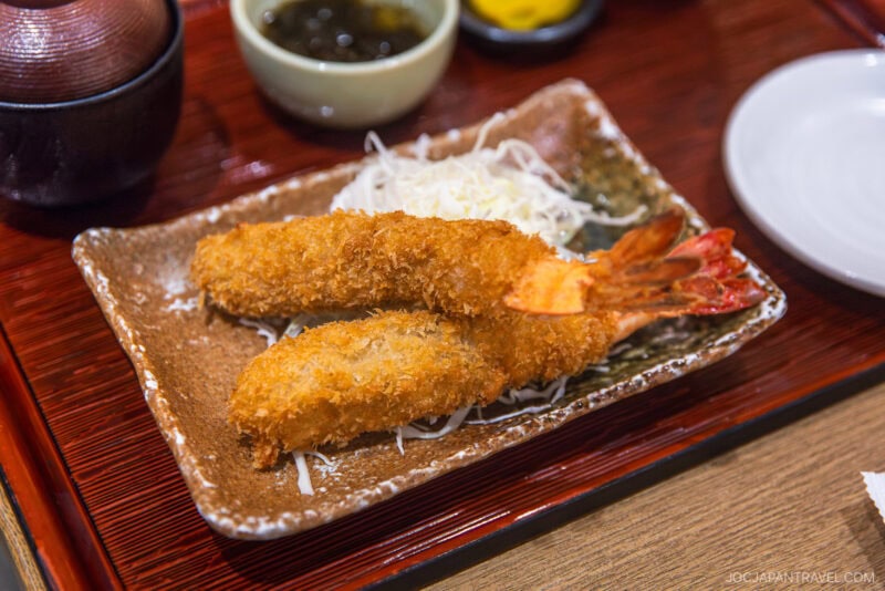 Two pieces of golden, crispy breaded fried shrimp on a brown rectangular plate, served on a bed of shredded cabbage with small bowls of dipping sauces—a must-try from any Nagoya food guide.