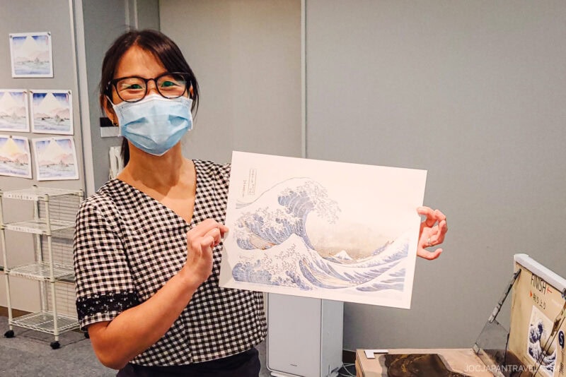 A woman wearing glasses and a face mask holds up a print of The Great Wave off Kanagawa in a room adorned with art, evoking the spirit of Japanese culture much like the historic Nakasendo Trail.