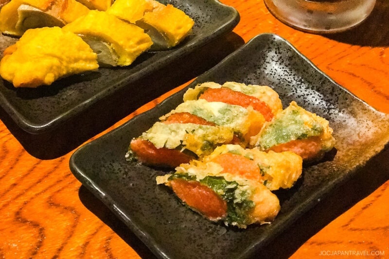 Close-up of tempura vegetables and fish served on black rectangular plates at Negibouzu Izakaya, with one plate containing yellow pieces and another featuring green and orange tempura items on a wooden table.