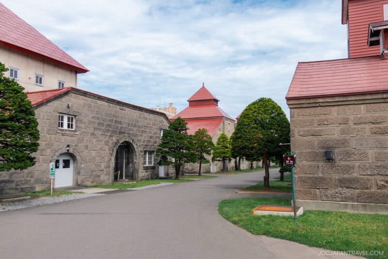 A paved pathway runs between stone buildings with red roofs and neatly trimmed trees under a partly cloudy sky at the historic Nikka Whisky Yoichi Distillery.