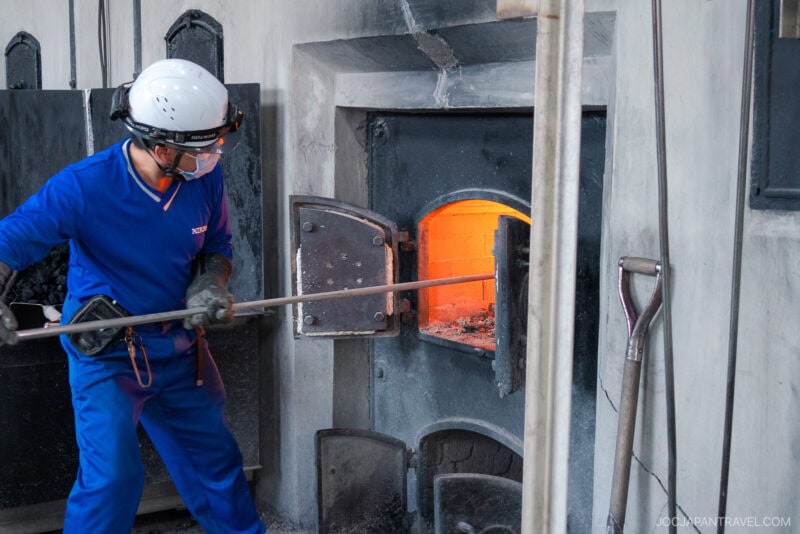 A worker in blue protective clothing, gloves, helmet, and mask uses a long metal tool to tend to a furnace with an open door at the Nikka Whisky Yoichi Distillery, revealing a glowing orange fire inside.