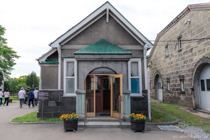 A small grey stone building with a green roof, arched entryway, double doors open, and flower pots at the entrance stands at Nikka Whisky Yoichi Distillery. People walk along a path nearby on a sunny day.