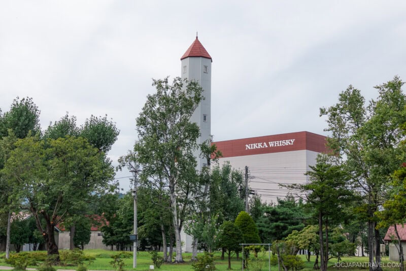 A tall building with a red roof stands near the Nikka Whisky Yoichi Distillery, surrounded by trees and greenery under a cloudy sky.