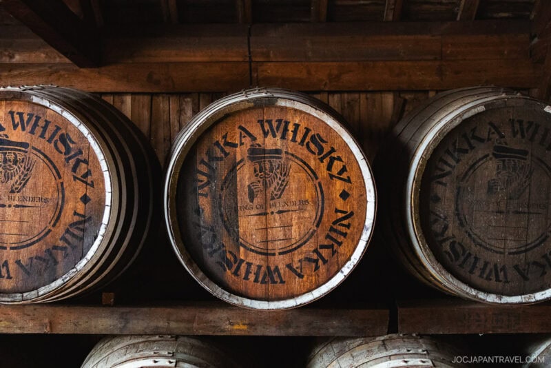 Three wooden barrels marked with NIKKA WHISKY and a logo rest on a wooden shelf inside the dimly lit Nikka Whisky Yoichi Distillery.