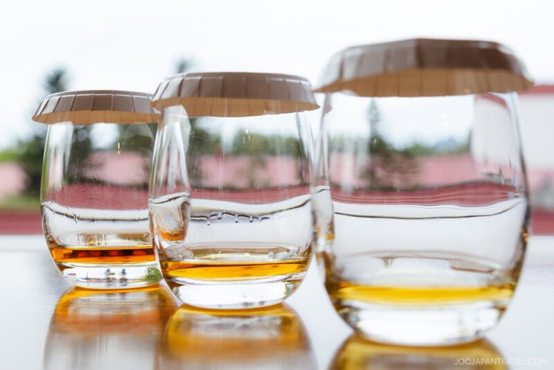 Three glasses with a small amount of amber liquid from Nikka Whisky Yoichi Distillery, each covered with a paper lid, are placed on a shiny surface with a blurred outdoor background.