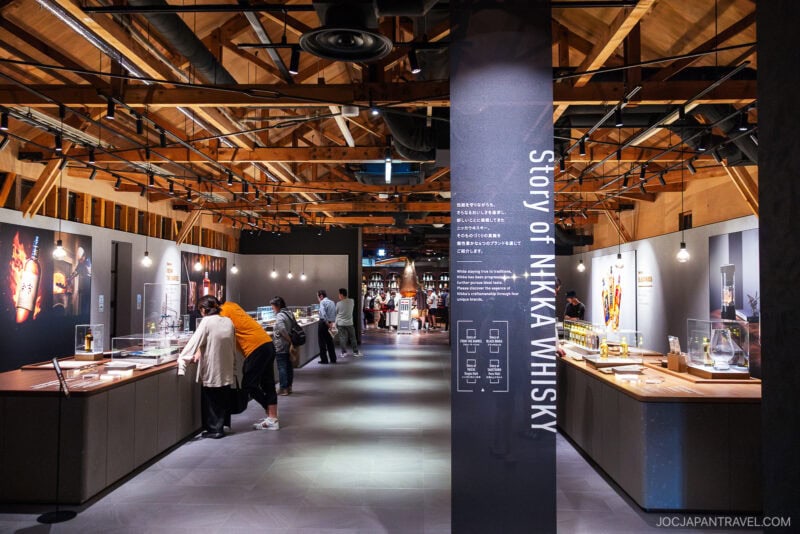 Visitors explore a modern whisky museum at Nikka Whisky Yoichi Distillery, with wooden ceilings and displays about the story of Nikka Whisky; exhibits and tasting stations line both sides of a central walkway.