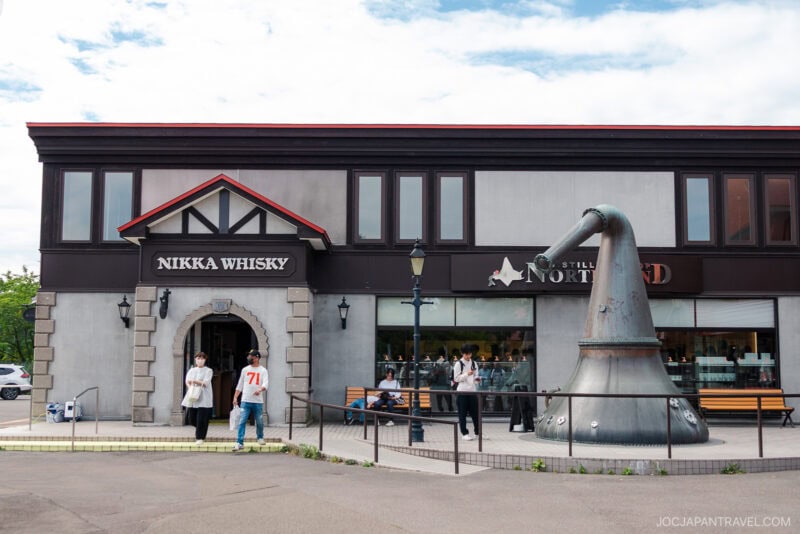 People stand outside the Nikka Whisky Yoichi Distillery building, which features a large distillation pot sculpture and a Still Northland sign. Some visitors are entering or sitting on a bench.