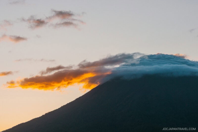 A mountain peak in Niseko autumn is partially covered by clouds, with a streak of orange sunlight illuminating the scene at sunset. The sky is pale, and the lower part of the mountain remains in shadow.