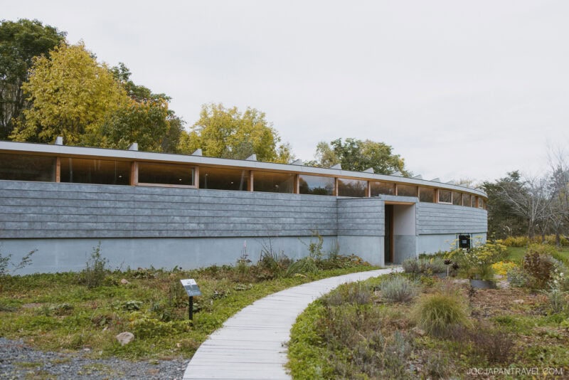 A curved, modern building with large windows and gray siding sits surrounded by greenery and wildflowers in a Niseko autumn setting, with a wooden pathway leading to its entrance. Trees with vibrant fall leaves are visible in the background.