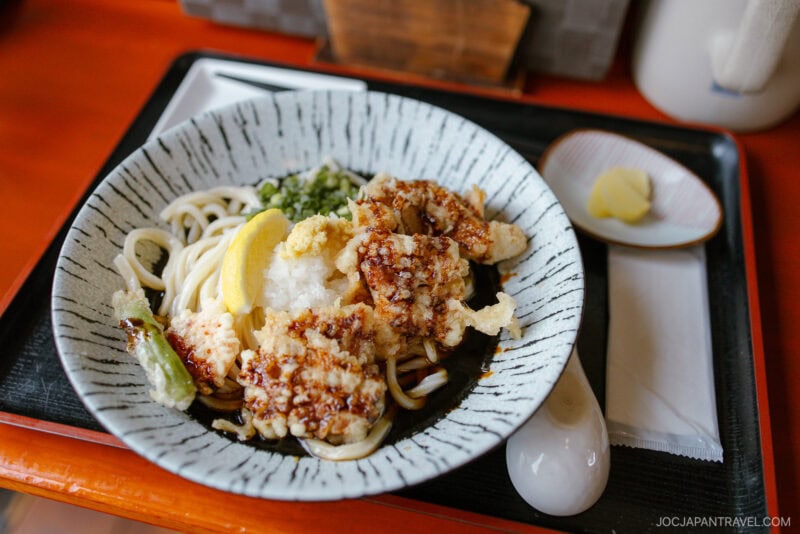 A bowl of udon noodles topped with tempura, grated radish, a lemon wedge, and green garnish evokes the cozy flavors of Niseko autumn, served on a tray with pickles, a ceramic spoon, and a napkin.