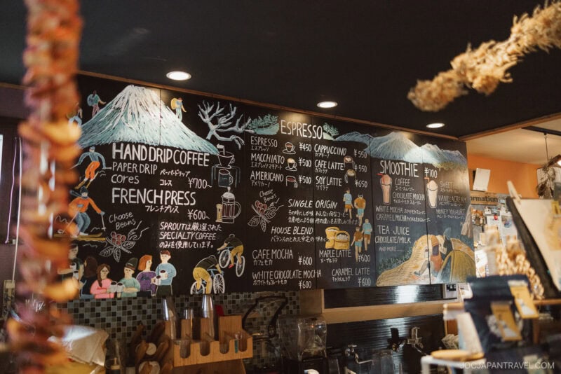 A chalkboard menu in a café shows colorful illustrations of Mount Fuji, coffee cups, and people enjoying Niseko autumn. Menu sections list hand drip coffee, French press, espresso drinks, and smoothies, with prices in Japanese yen.