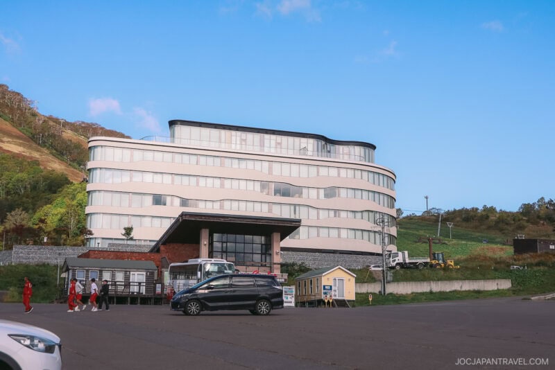 A modern, multi-story white building with curved glass windows stands by a hillside under a clear blue sky in Niseko autumn. Several cars and small buildings are in the parking lot, and a group of people walk nearby.