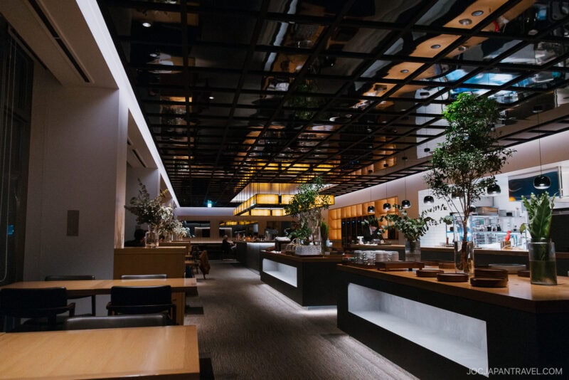 Modern restaurant interior with wooden tables, chairs, and large green plants evokes the cozy ambiance of a Niseko autumn. The reflective grid ceiling and warm lighting add charm, while counters with plates and utensils line the center of the space.