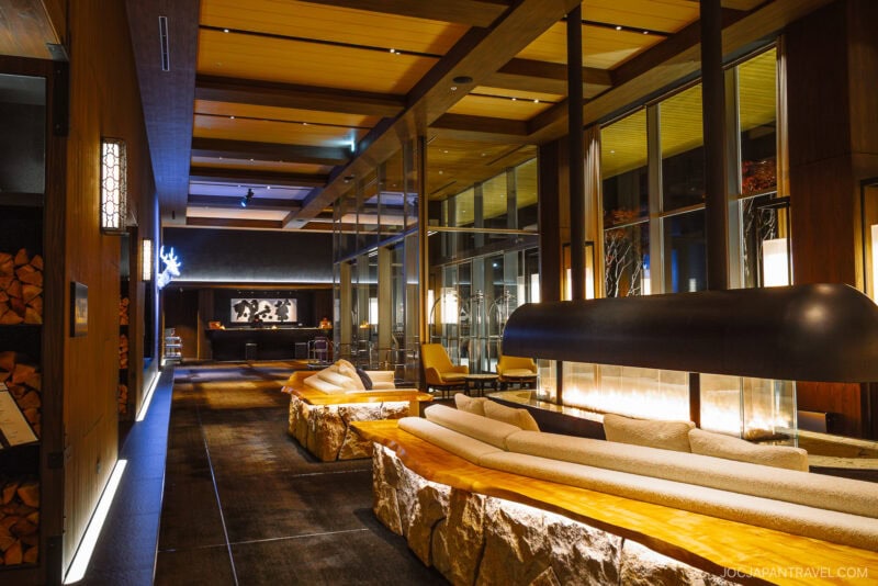Modern hotel lobby with warm lighting, wooden accents, and large glass windows. Stone benches with wooden seats surround a central fireplace. Elegant décor creates a cozy atmosphere, perfect for enjoying Niseko autumn views.