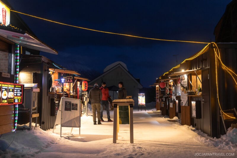 Three people stand talking in the middle of a snow-covered outdoor market at night, surrounded by food stalls lit with warm lights and colorful signs—a perfect scene for enjoying Niseko winter activities. Snow piles are visible along the walkway.