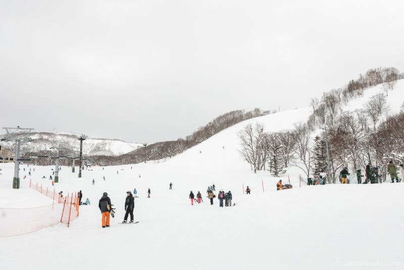 People enjoy skiing and snowboarding on a wide, snowy ski slope surrounded by hills and trees on a cloudy day, with ski lifts and skiers visible in the background—perfect for experiencing Niseko winter activities.
