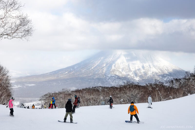 Snowboarders and skiers descend a snowy slope with a large, snow-capped mountain in the background, partially obscured by clouds. Trees line the edge of the slope under a cloudy sky, capturing the excitement of Niseko winter activities.