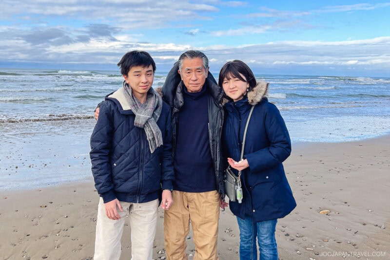 Three people stand close together and smile on a sandy beach with the ocean and blue sky of the Noto Peninsula in the background. They wear jackets and scarves, suggesting cool weather.