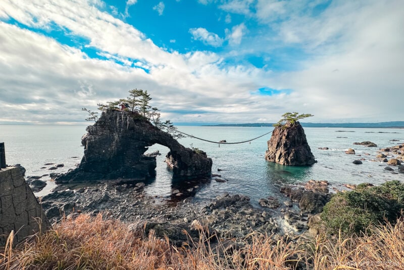 Two rocky islets connected by a sacred rope, with small trees atop, rise from the calm blue sea off the Noto Peninsula. Scattered rocks and grassy plants line the scene under a partly cloudy sky.