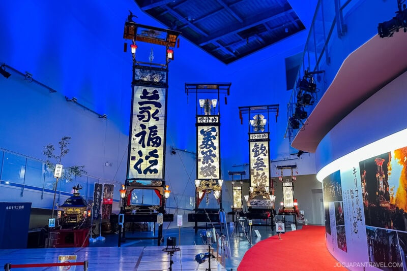 Large traditional Japanese festival floats from the Noto Peninsula with tall banners are displayed in a brightly lit indoor museum. The space features blue lighting, a red carpet, and curved white walls adorned with photos and exhibits.