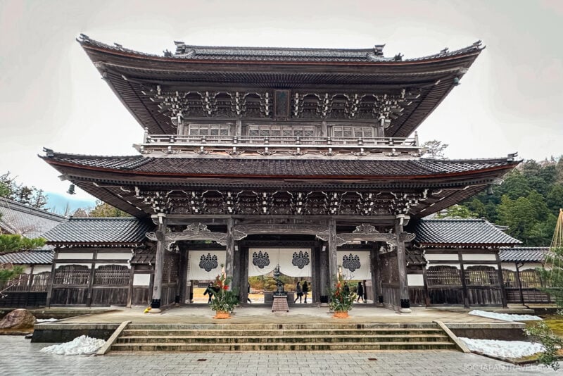 A traditional Japanese wooden temple with ornate carvings and a two-tiered roof stands on the Noto Peninsula, set against a cloudy sky. Small plants and patches of snow are visible near the entrance.