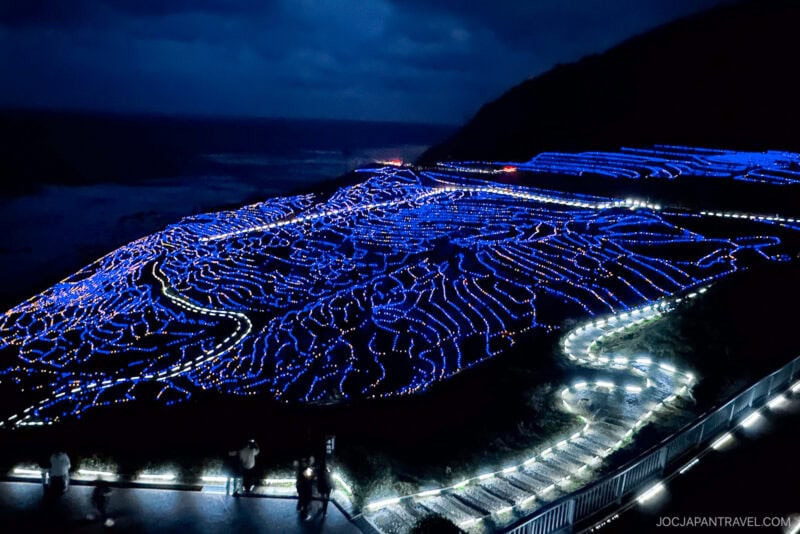 Terraced rice fields on the Noto Peninsula glow with blue LED lights at night, creating mesmerizing patterns across the hillside. Pathways shine with white lights as a few people observe the illuminated landscape under a dark, cloudy sky.