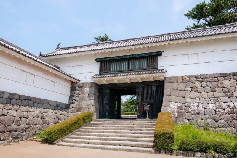 Stone steps lead to the traditional Odawara Castle gate with white plaster walls, a tiled roof, and dark wooden doors, surrounded by historic stone walls and lush greenery under a clear blue sky.